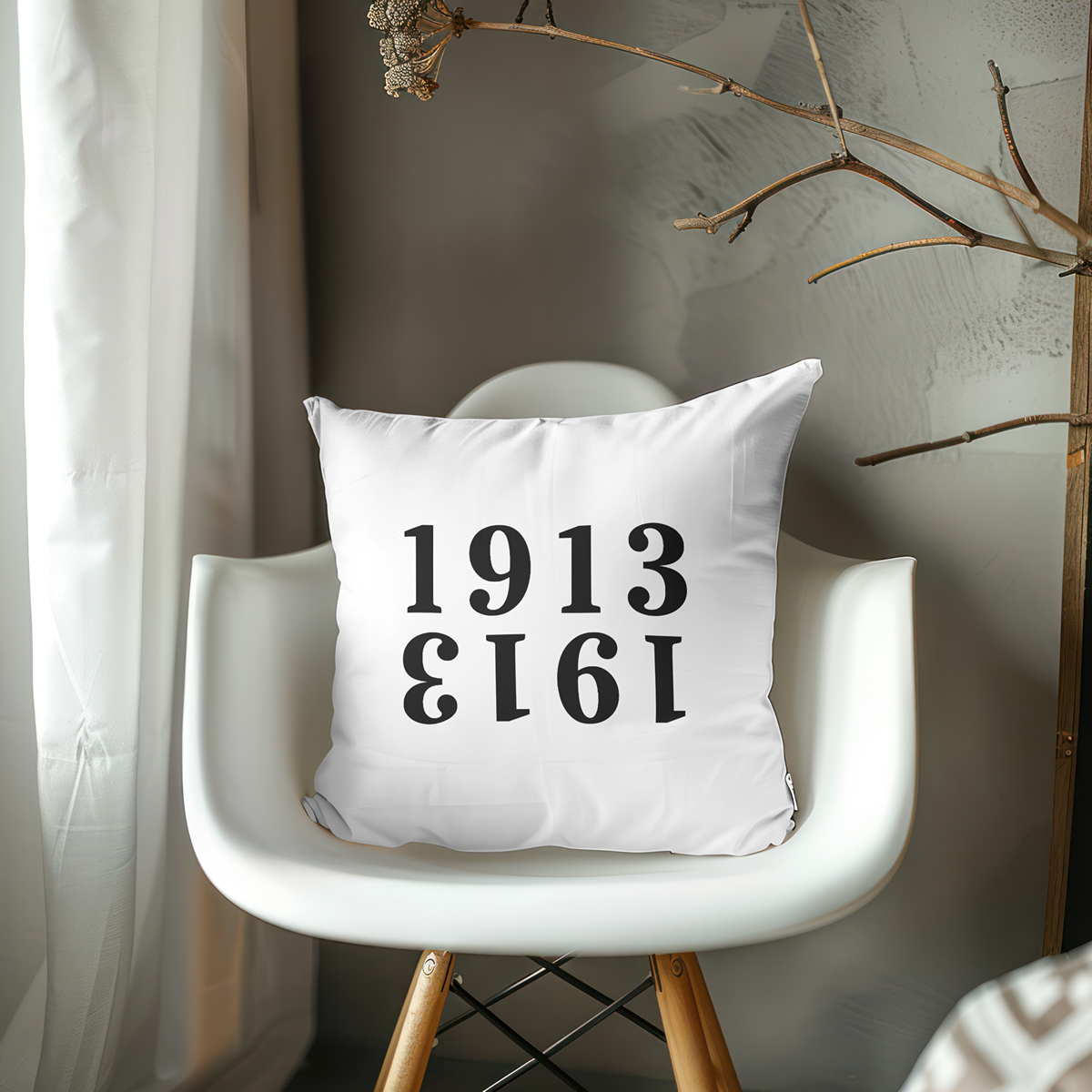 White pillow from The Orange Habit with black text '1913' on a white chair against a neutral wall.