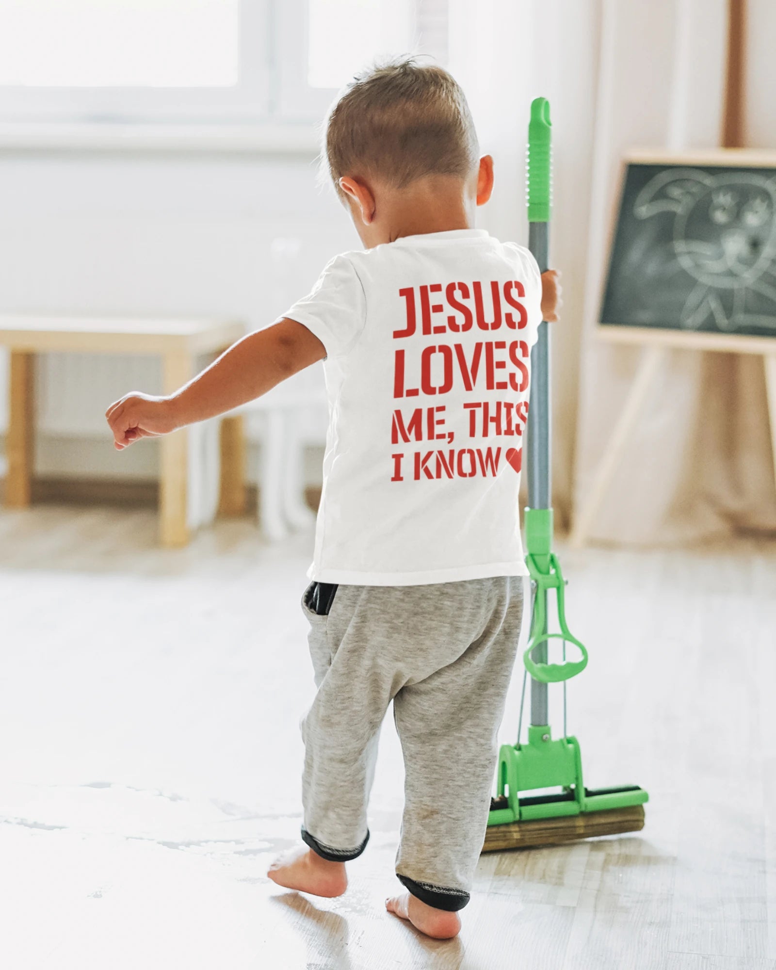 Child wearing a shirt with 'Jesus loves me, this I know' while holding a mop.