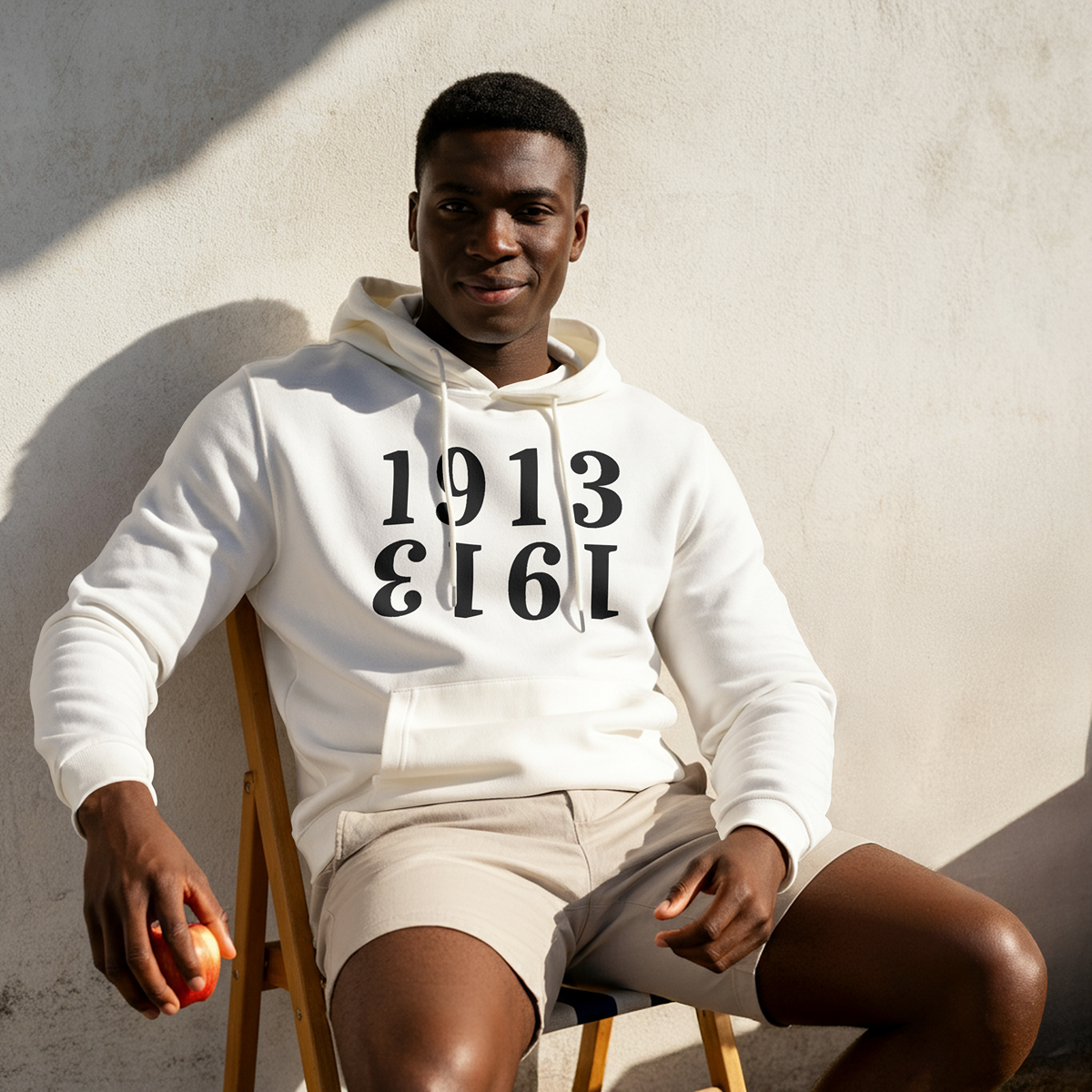 Man wearing a white hoodie from The Orange Habit with '1913' printed on it, sitting against a light-colored wall.