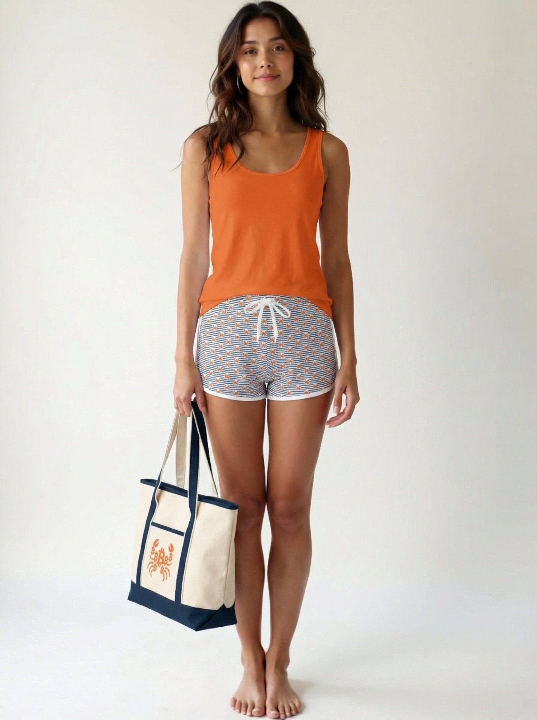 Woman wearing an orange tank top and bitcoin crab patterned shorts, holding a tote bag on a white background | SVRN Society
