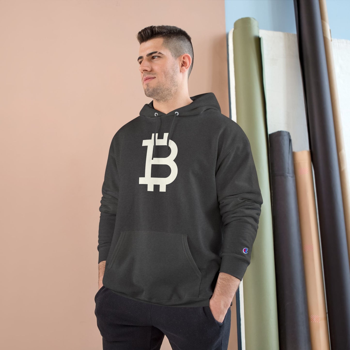 Man wearing a (Charcoal Grey) hoodie with a Bitcoin logo against a beige wall.
