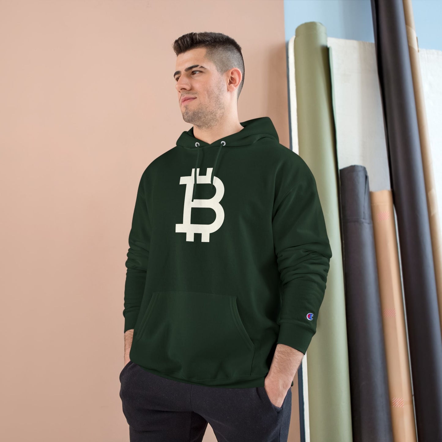 Man wearing a (Dark Green) hoodie with a Bitcoin logo against a beige wall.