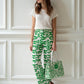 Woman wearing green patterned fix the money fix the world Bitcoin pants and holding a matching bag against a white paneled wall.