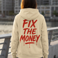 Rear view of the apricot Fix The Money hoodie with red brush-lettered back graphic beside the waterfront.