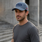 Model wearing the Denim Not A FED Sunwashed Cap with navy front embroidery beside gray concrete buildings.
