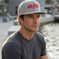 Model wearing the Heather Grey Kayfabe snapback with red script embroidery and green undervisor beside the waterfront.