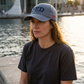 Model seated by the waterfront wearing the Denim Not A FED Sunwashed Cap with navy FED lettering and red side embroidery.