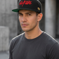 Model wearing the Black Kayfabe snapback with red script and gold theater-mask embroidery beside concrete buildings.