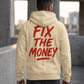 Model wearing the apricot Fix The Money zip-up hoodie, showing the red back graphic beside concrete buildings.
