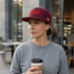 Model holding coffee outside a cafe in the Maroon Kayfabe snapback with red front embroidery and a flat brim.