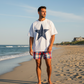 Man standing on a beach wearing a white t-shirt with a blue star and red plaid shorts.