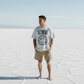 Man wearing a t-shirt with a Satoshi smiley face design and text standing on a salt flat. - SVRN Society