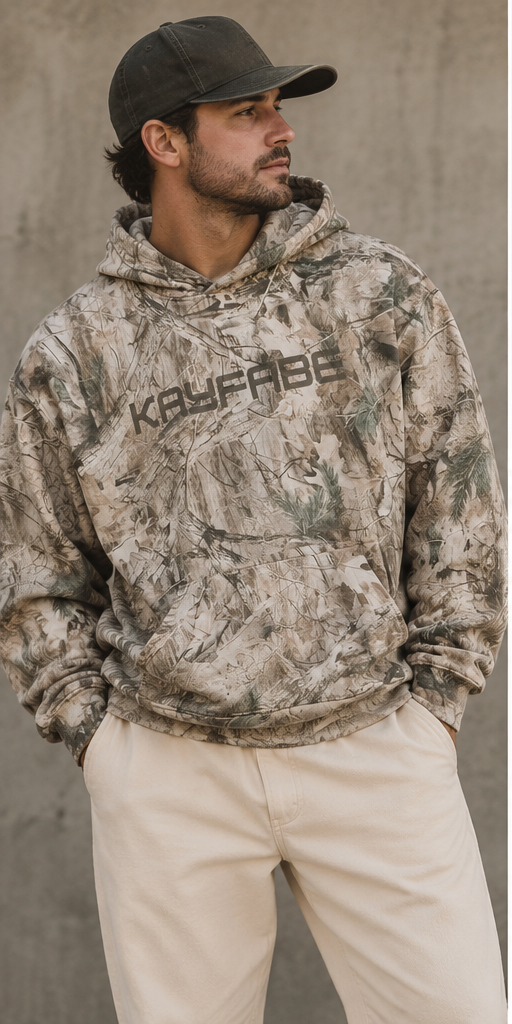 Man in a cap wearing the Kayfabe tree camo oversized fleece hoodie with cream pants