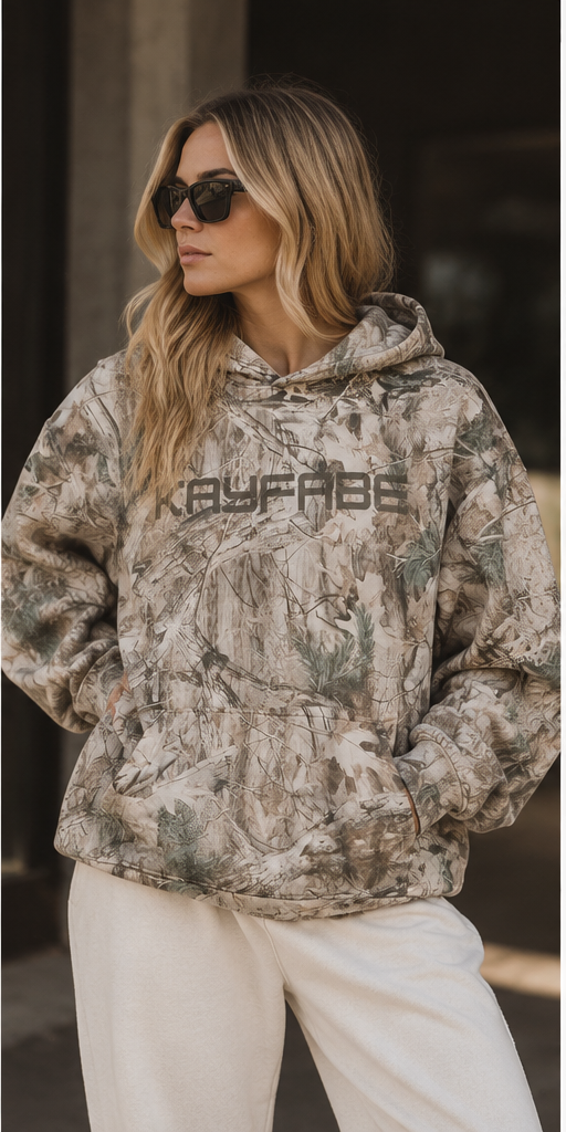 Woman in sunglasses wearing the Kayfabe tree camo oversized fleece hoodie with cream pants