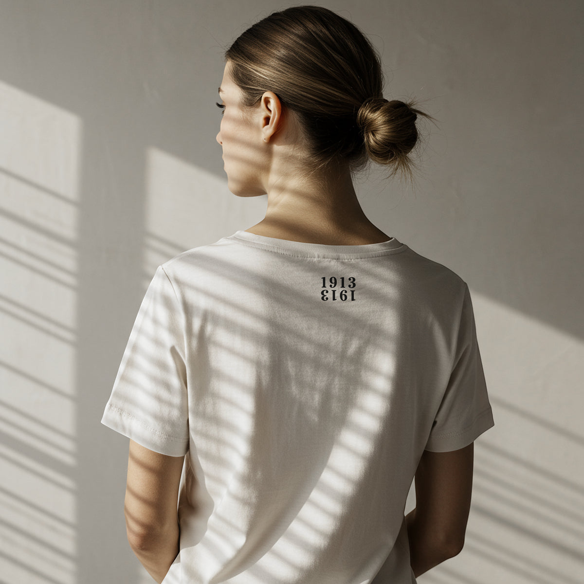 woman wearing a white t-shirt from The Orange Habit Bitcoin Clothing Store with 1913 on the back, standing in front of a sunlit wall.
